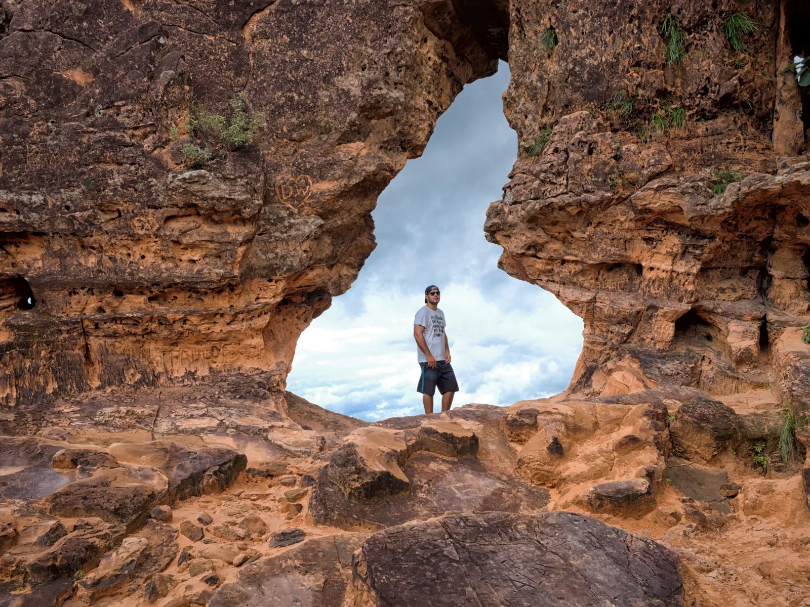 A man standing inside a large keyhole-shaped opening in a wall of weathered orange sandstone, cloudy sky behind him