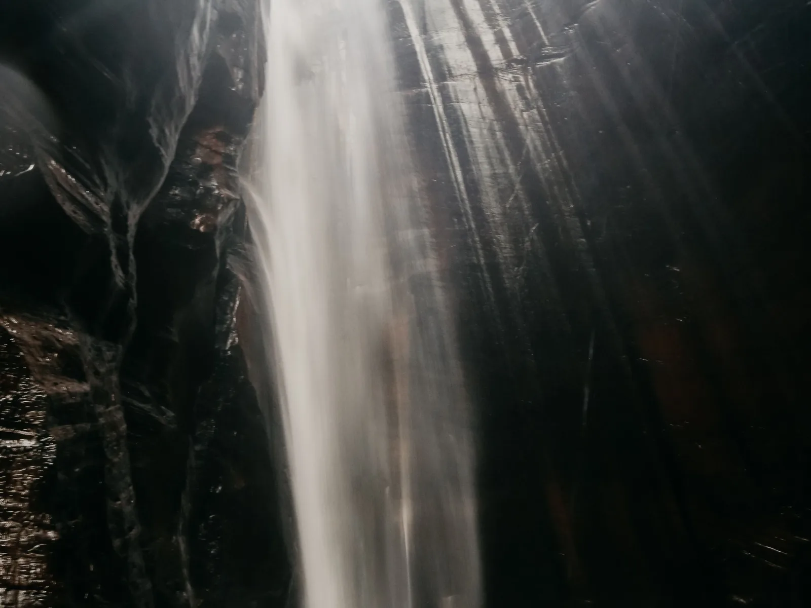 A tall, thin waterfall falling down a dark canyon wall into a pool, lit by shafts of sunlight coming through the gap above