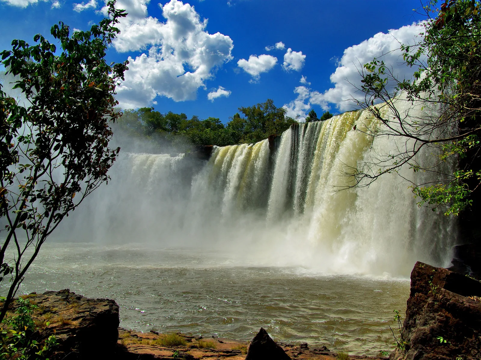 A wide waterfall falling in many white streams off a long ledge into a brown pool, with trees framing the view and blue sky full of clouds above