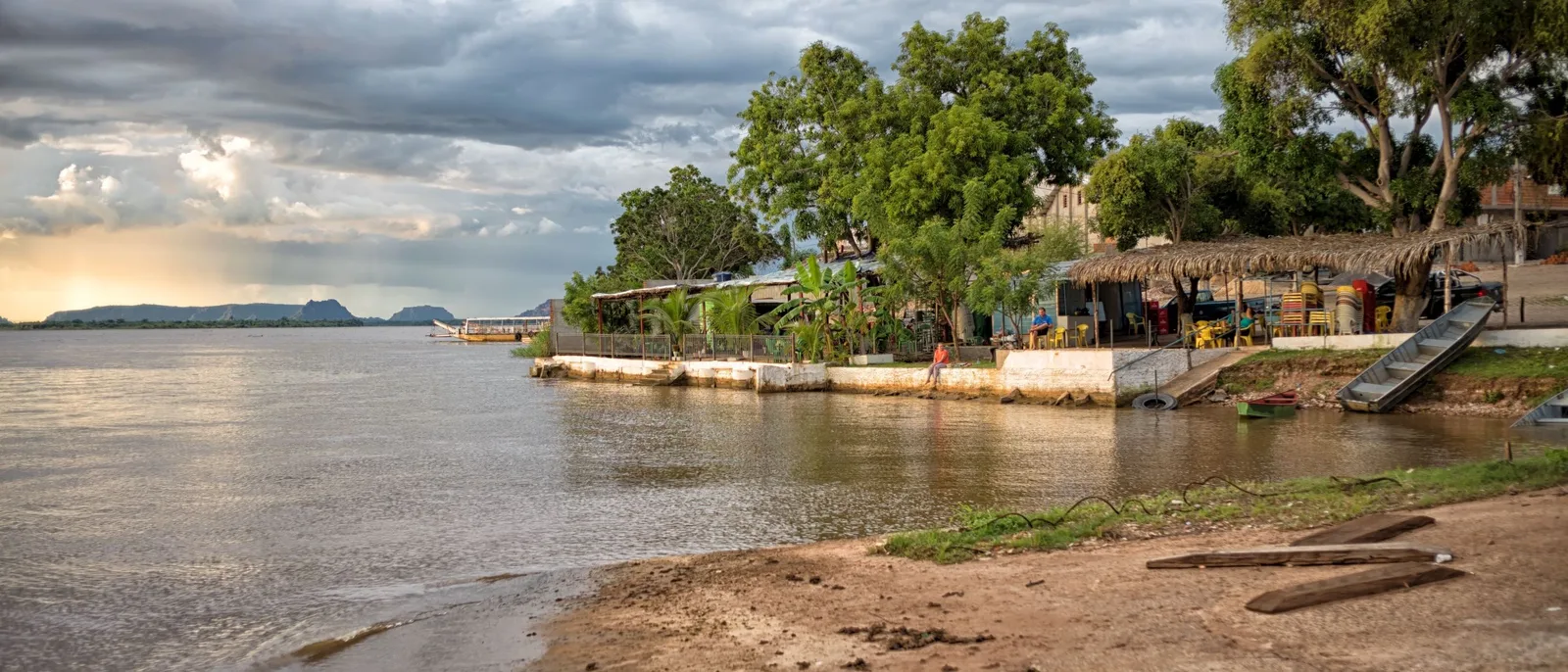 The Carolina riverfront under a cloudy sky, trees and thatched kiosks along a low embankment, a sandy bank and the wide Rio Tocantins in front