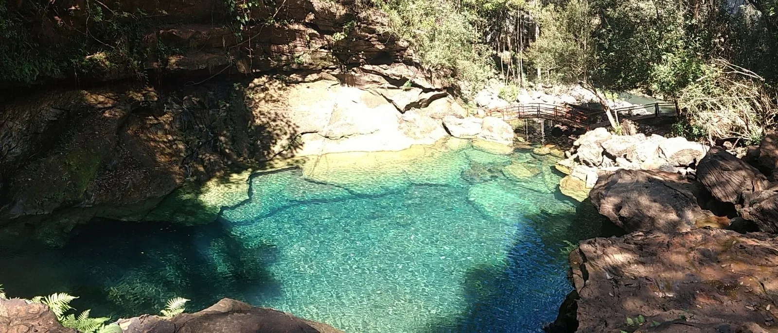 A wide view of a turquoise pool among sunlit boulders and trees in a rocky gorge at Poço Azul