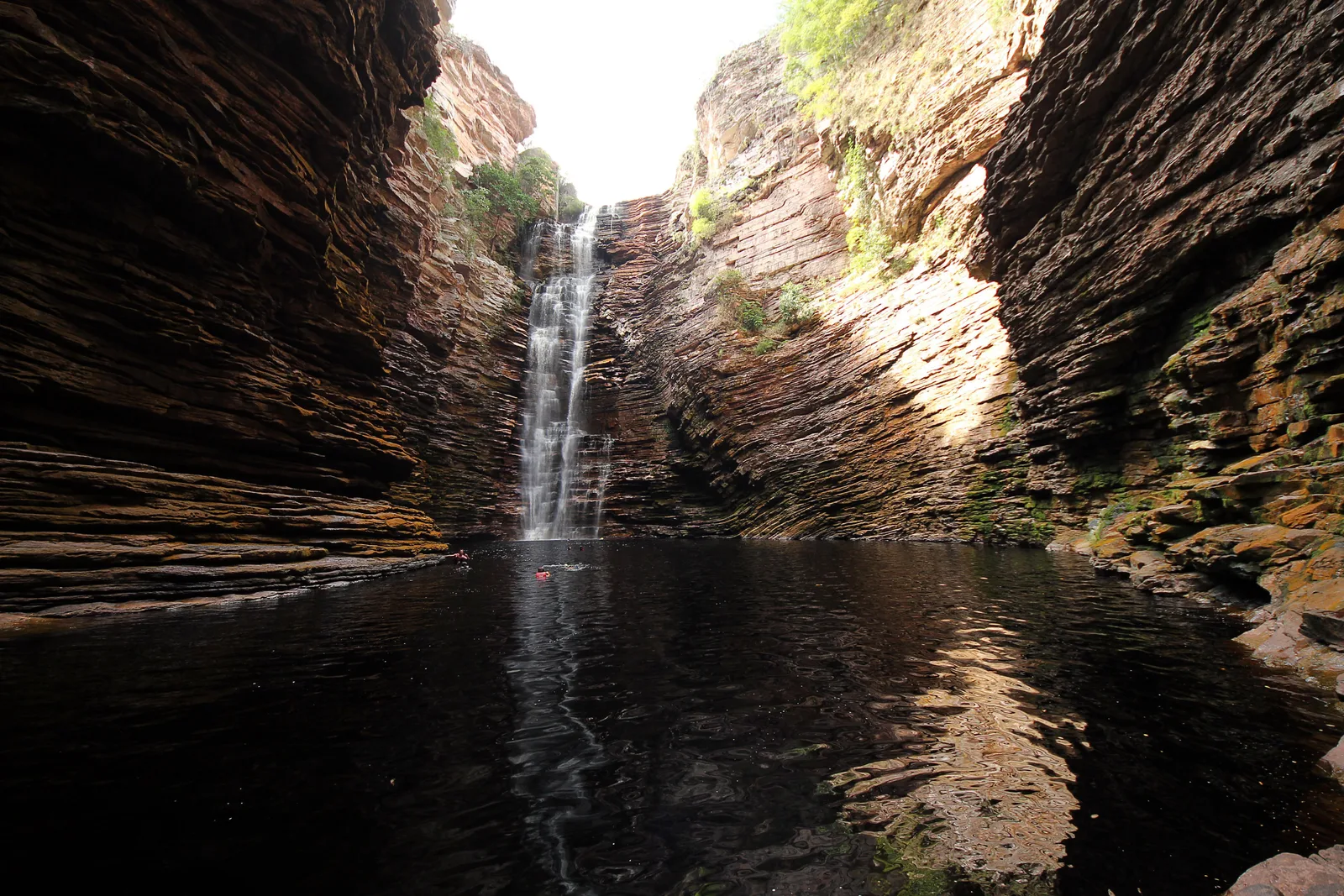 A tall waterfall dropping into a still dark pool at the head of a narrow canyon of banded rock