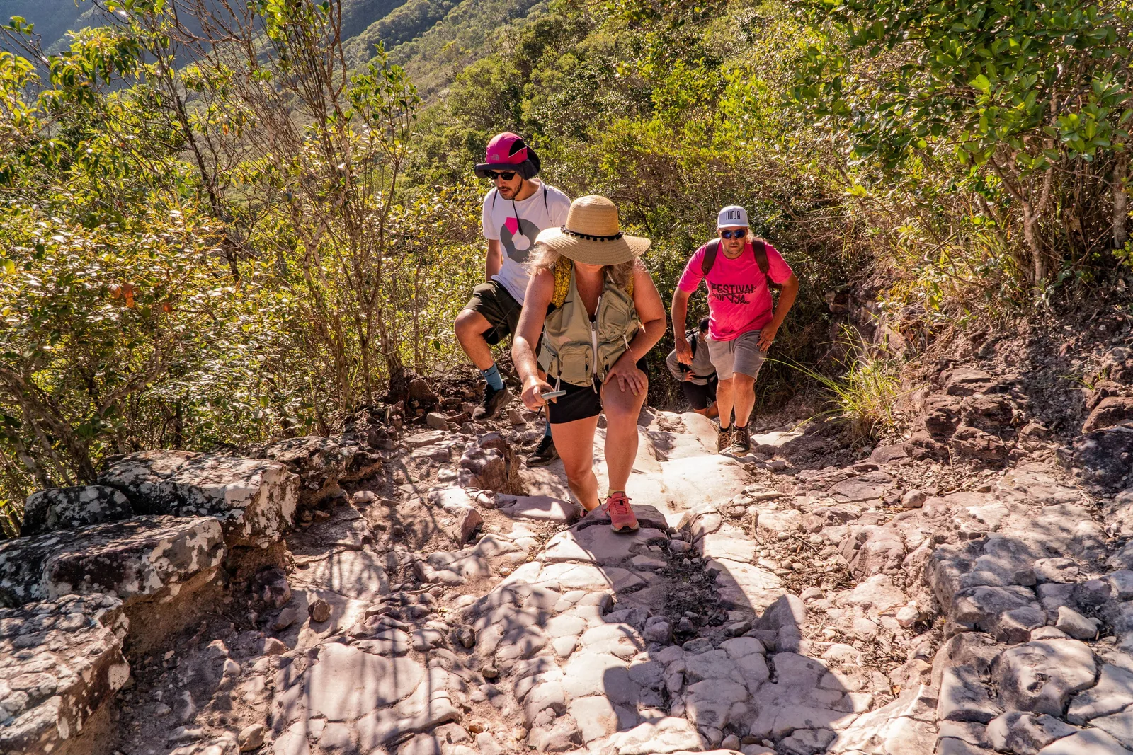 Four walkers, one in a straw sun hat and two in caps, on a rocky path through low scrub on the trail to Cachoeira da Fumaça