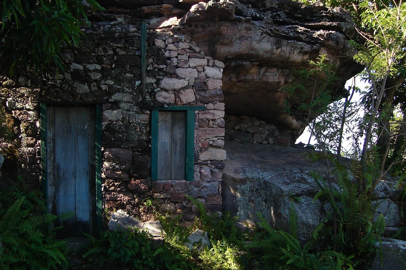 A stone house at Igatu with two weathered blue-green doors, built up against a rock outcrop with vegetation growing over it