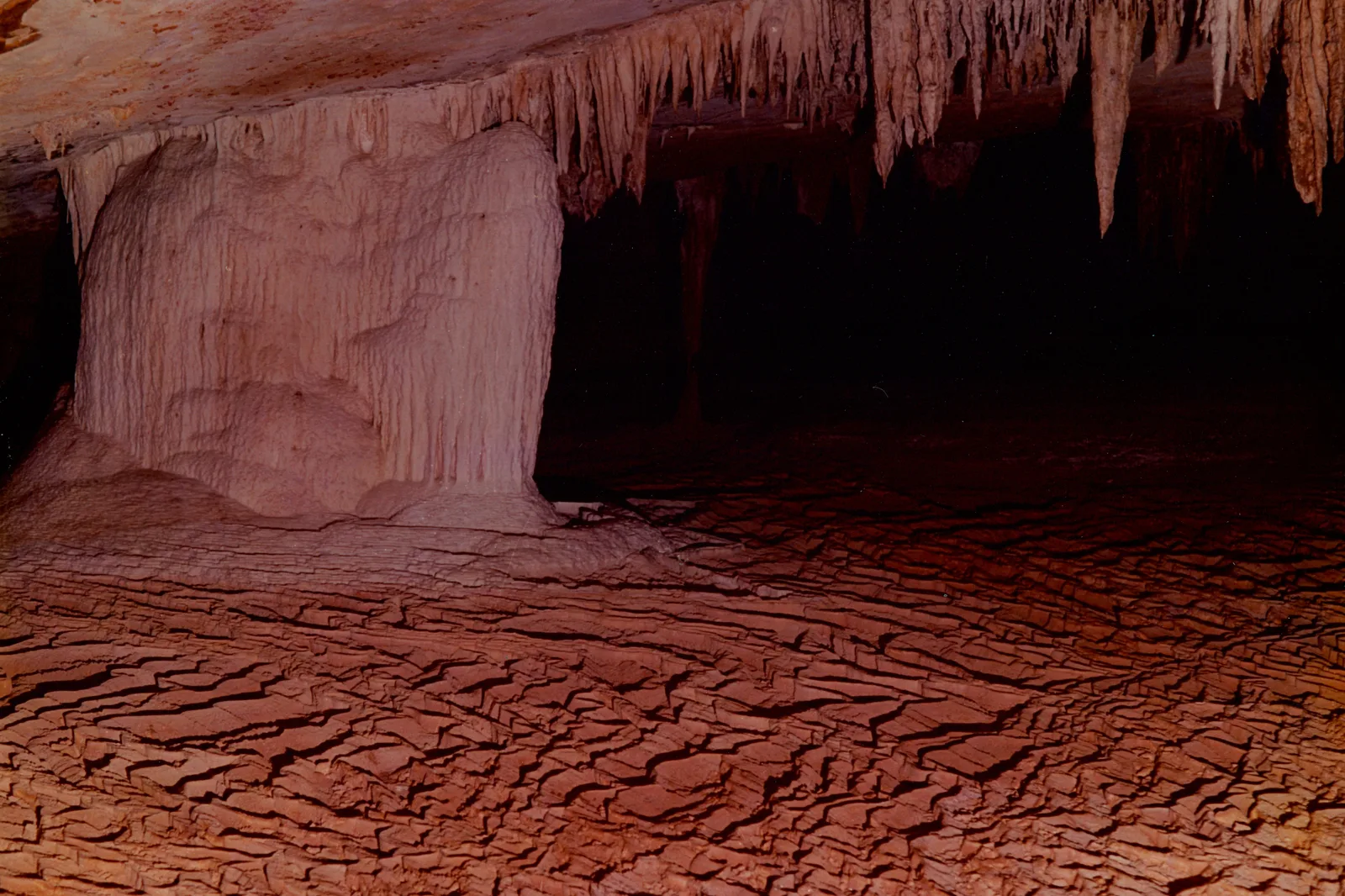 A vast cave gallery lit red-orange, stalactites hanging from the roof and a floor of cracked dried mud below