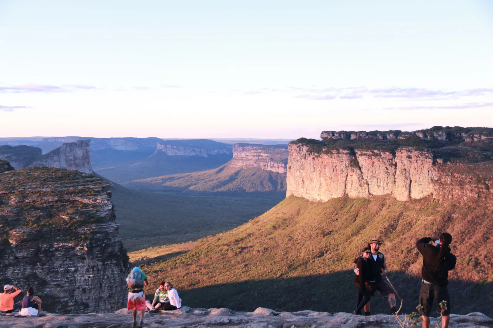 Visitors standing and sitting on the summit of Morro do Pai Inácio, photographing a broad red-lit mesa across the valley