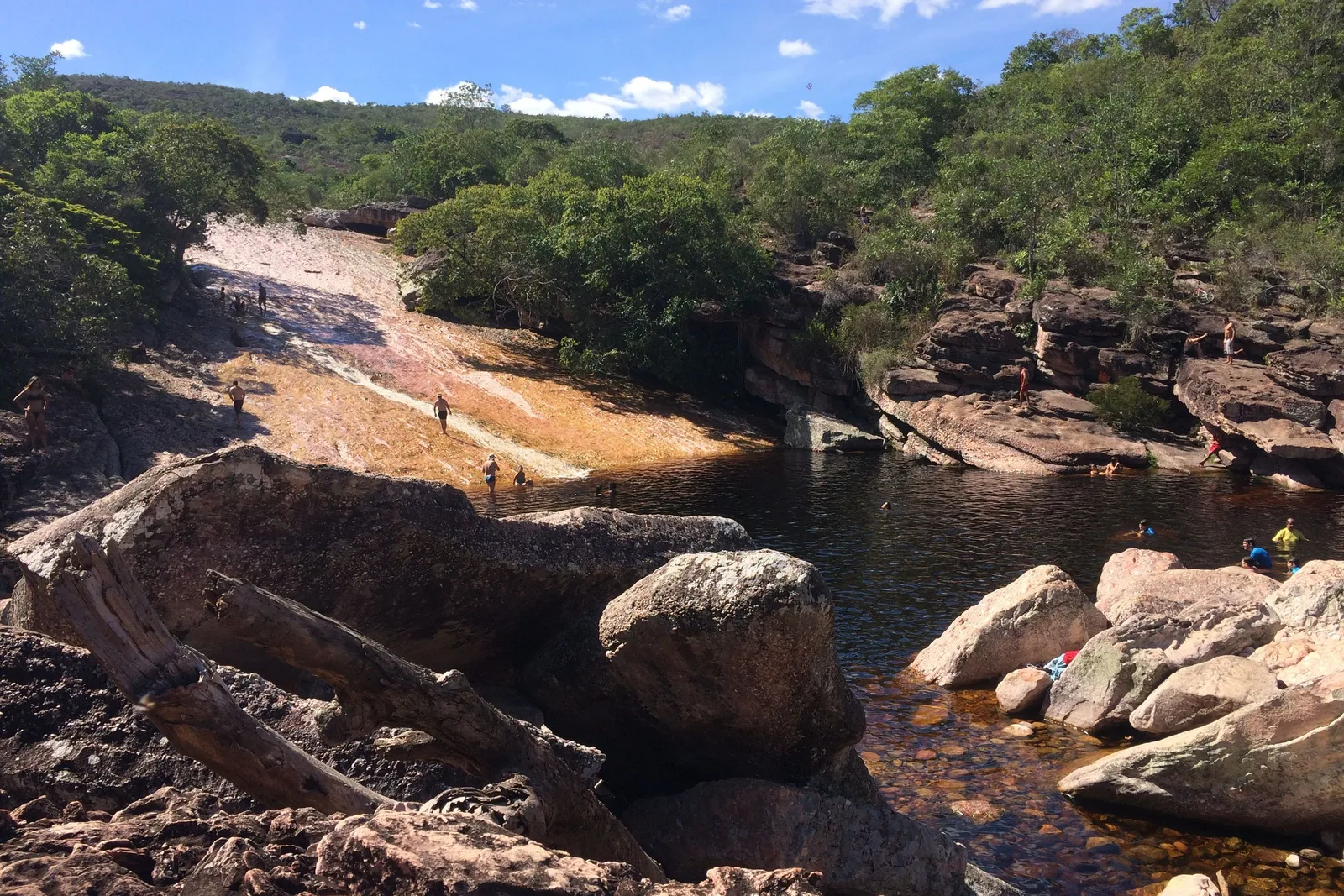 The river at Ribeirão do Meio running down a broad slope of smooth pale rock into a dark pool among boulders, with people small at the top of it