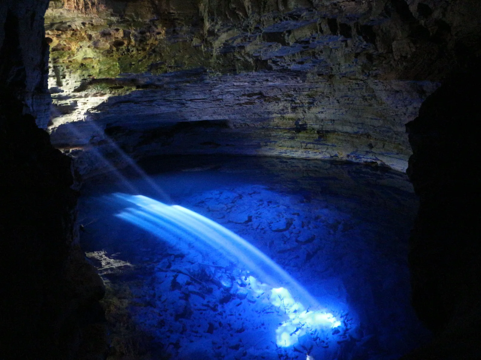 A shaft of sunlight striking the surface of a deep flooded cave pool and lighting the water electric blue