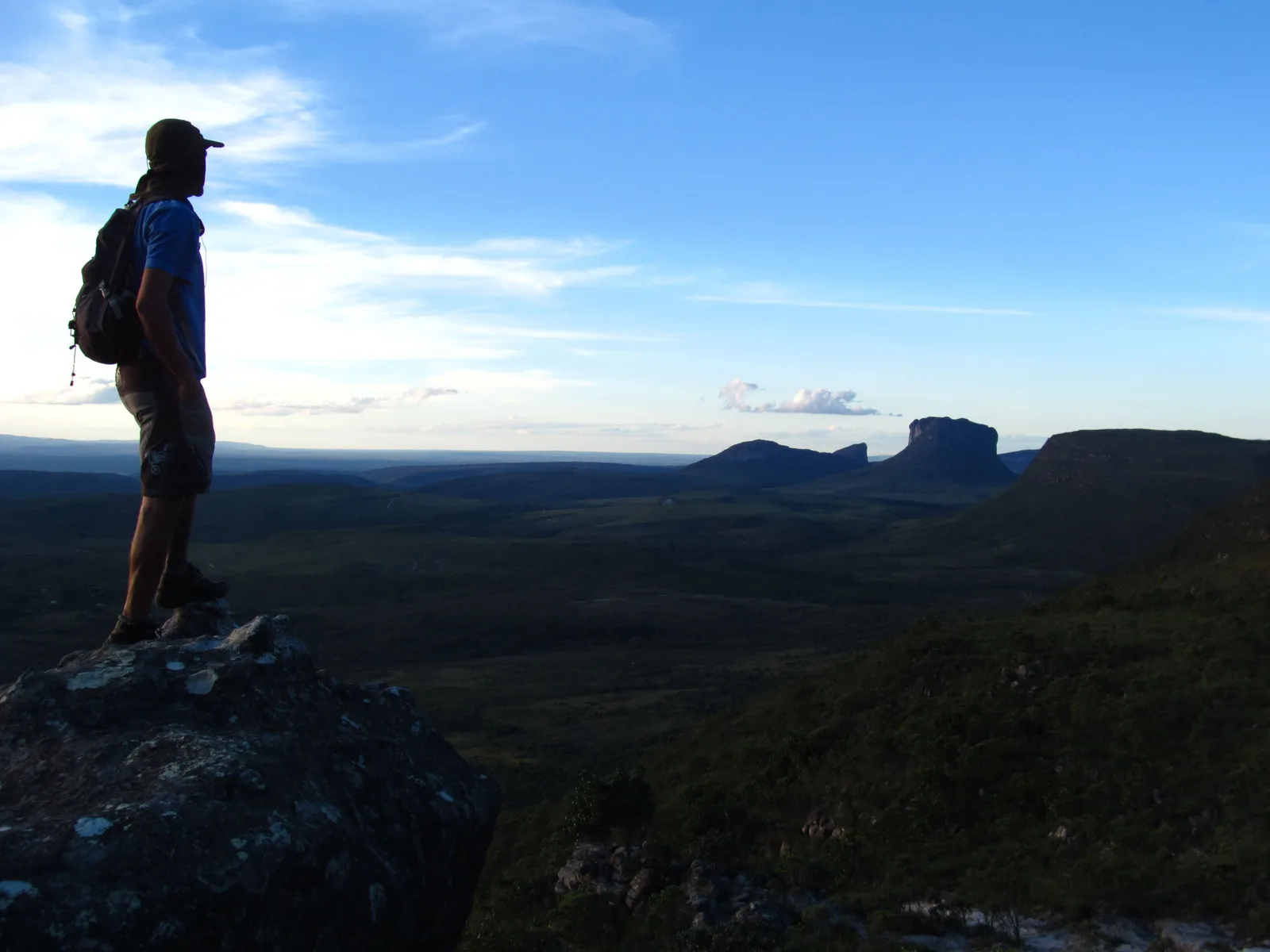 A walker with a backpack standing on a rock outcrop looking out over low cerrado scrub toward a distant flat-topped mountain