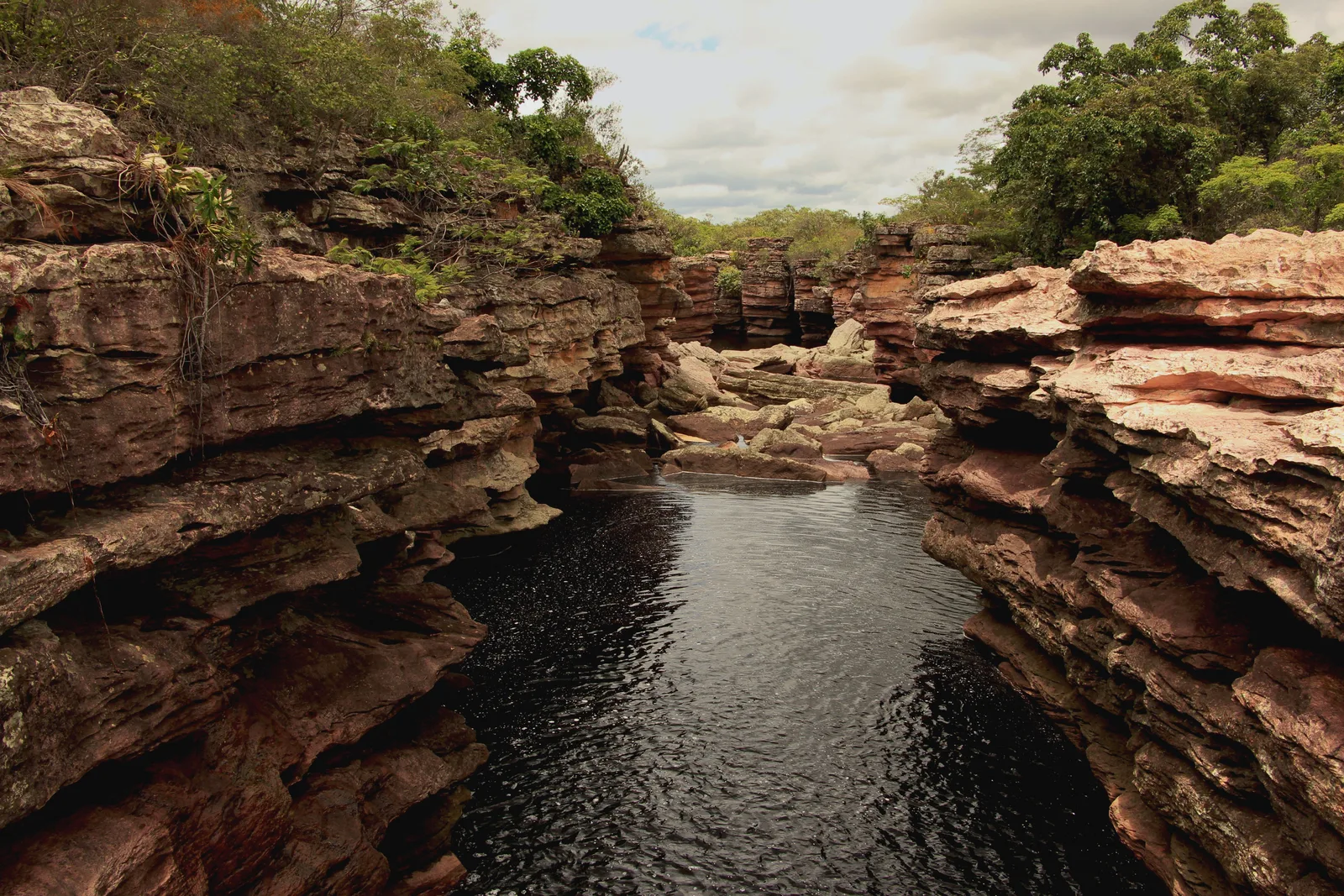 A dark river pool held between banded rock walls on the approach to Cachoeira do Buracão, trees along the top