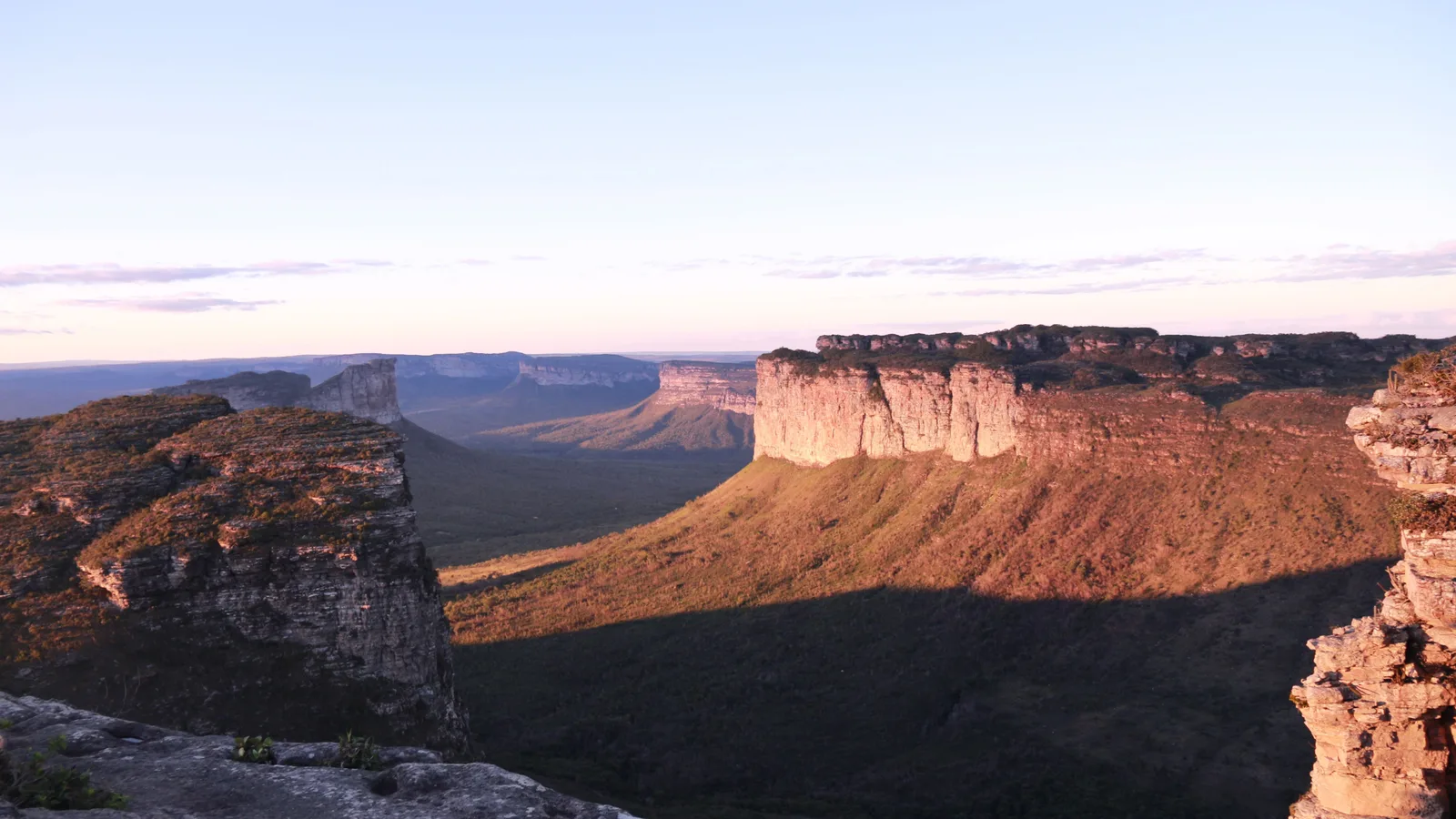 Flat-topped sandstone mountains stepping away across a wide green canyon in warm late light, one broad red-lit mesa standing closest to the camera