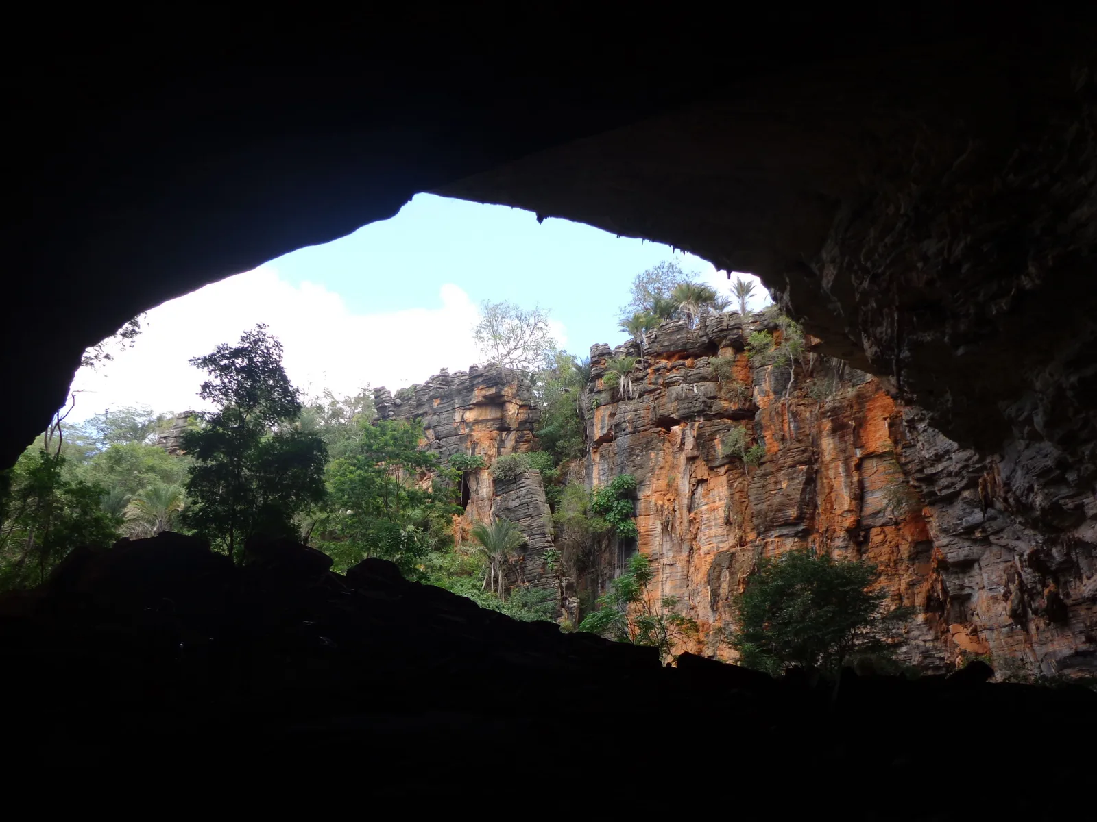 Looking out of the wide natural mouth of the Lapa Doce cave to a red rock face and trees in daylight