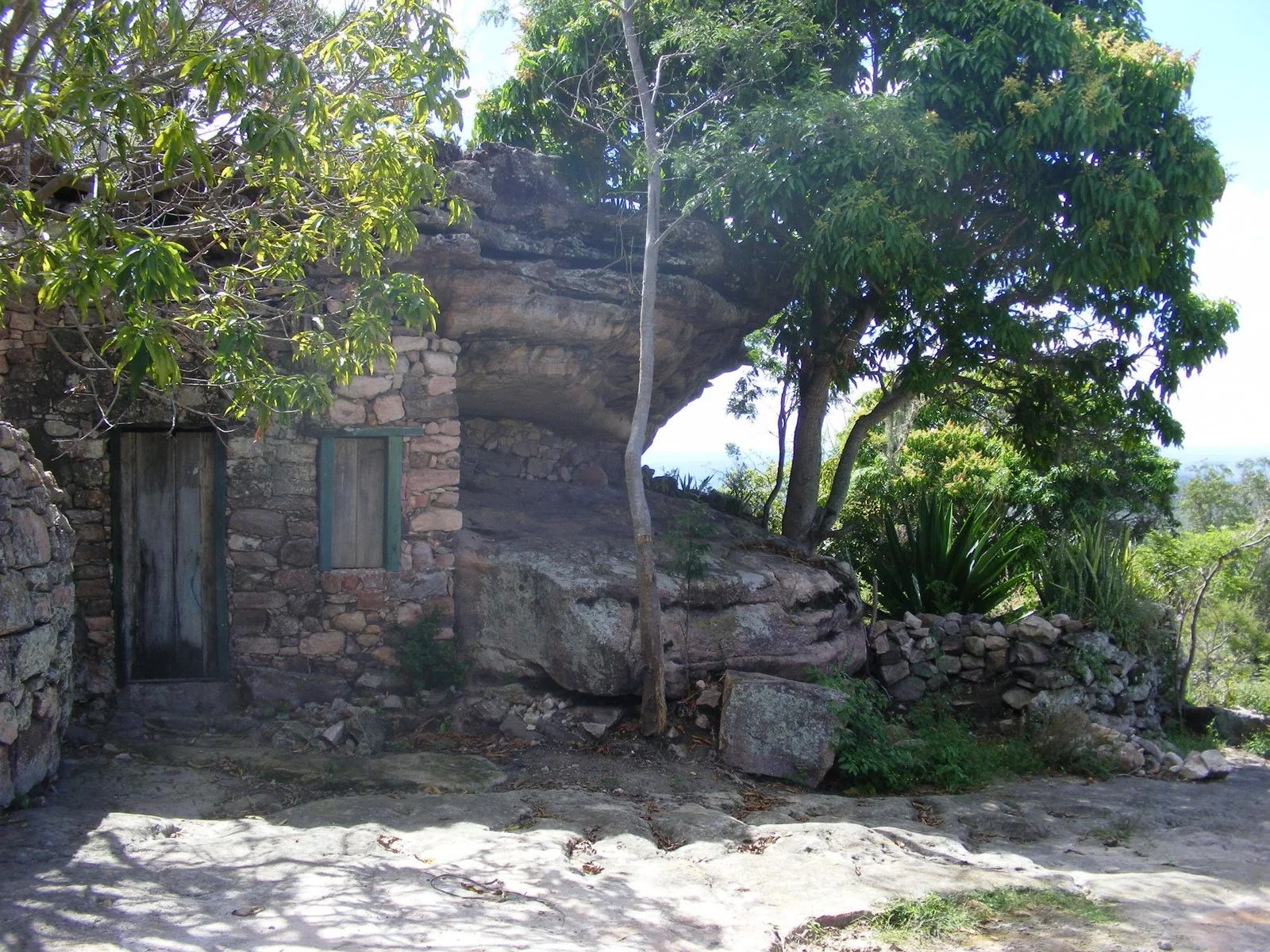 The Casa na Pedra at Igatu, a stone house roofed by an overhanging boulder, its green door intact, a tree standing in the open ground in front of it