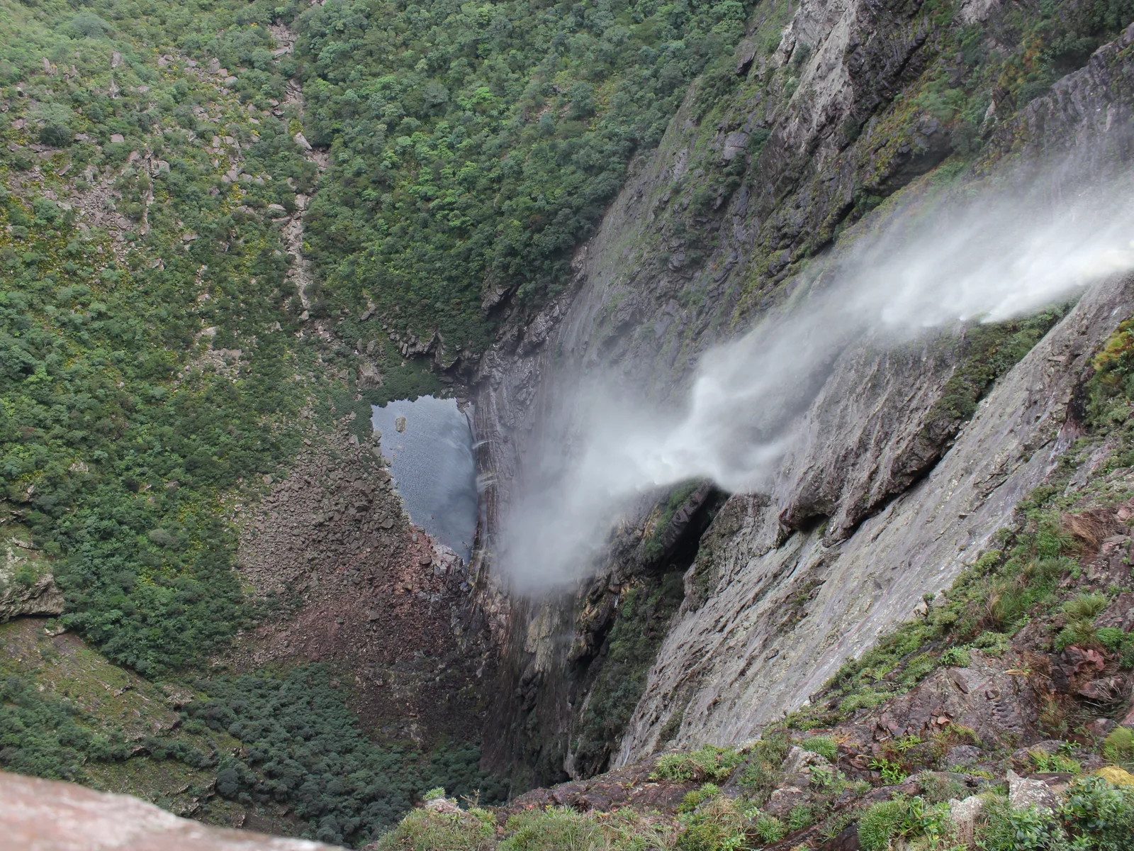Cachoeira da Fumaça seen straight down from the clifftop, the water blowing out into spray against a gray rock wall on the right, the wooded canyon floor far below
