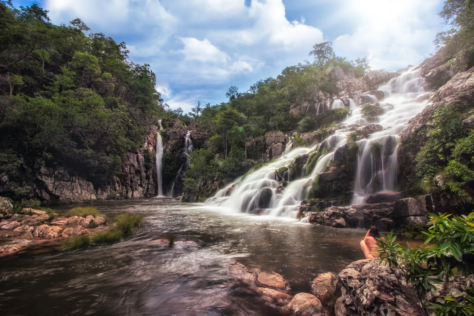 Water cascading in white streams down a rocky slope into a pool, with a second thin fall at the back of a wooded gorge