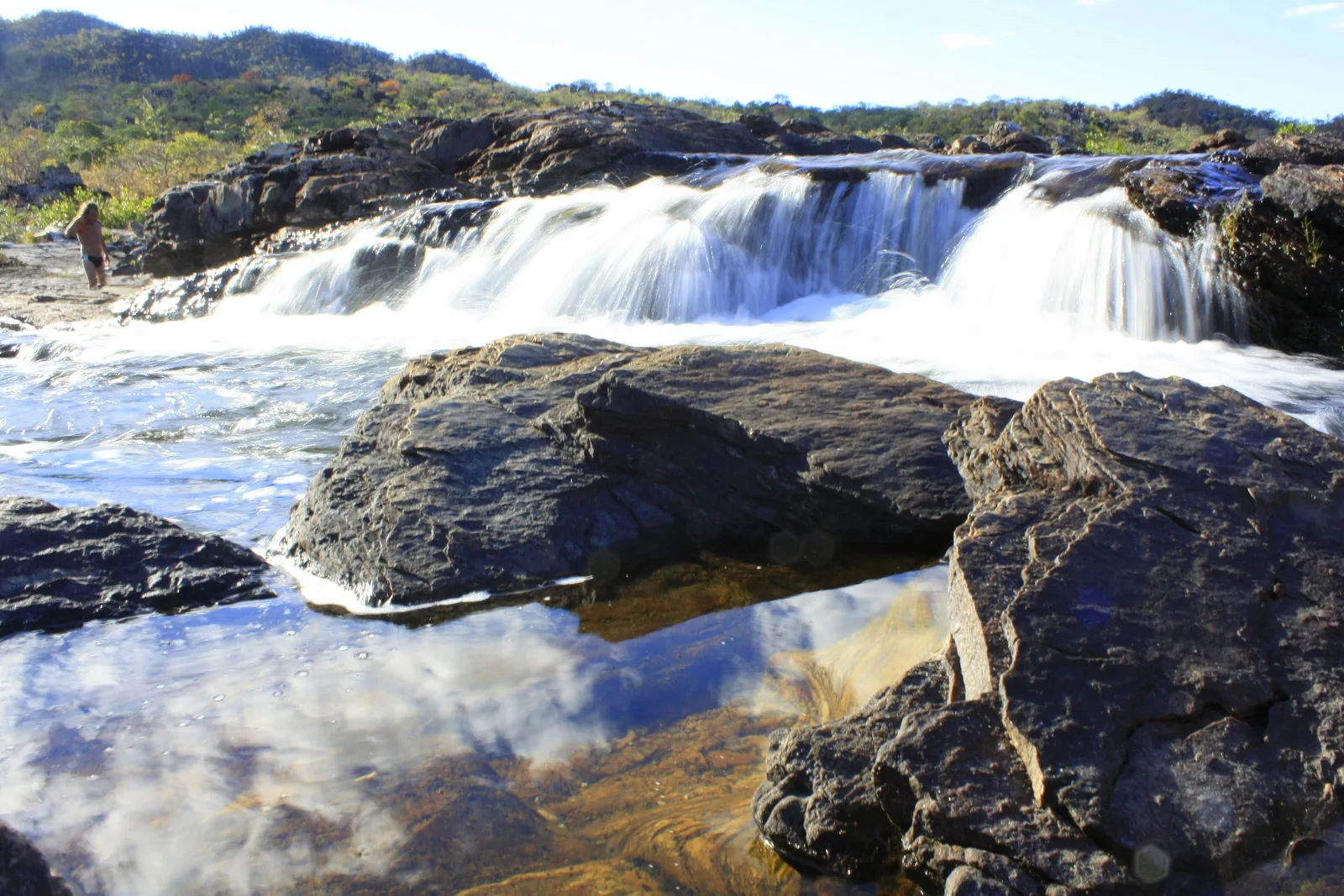 Water spilling in white sheets over a dark rock ledge, with a still pool in front reflecting the sky and clouds