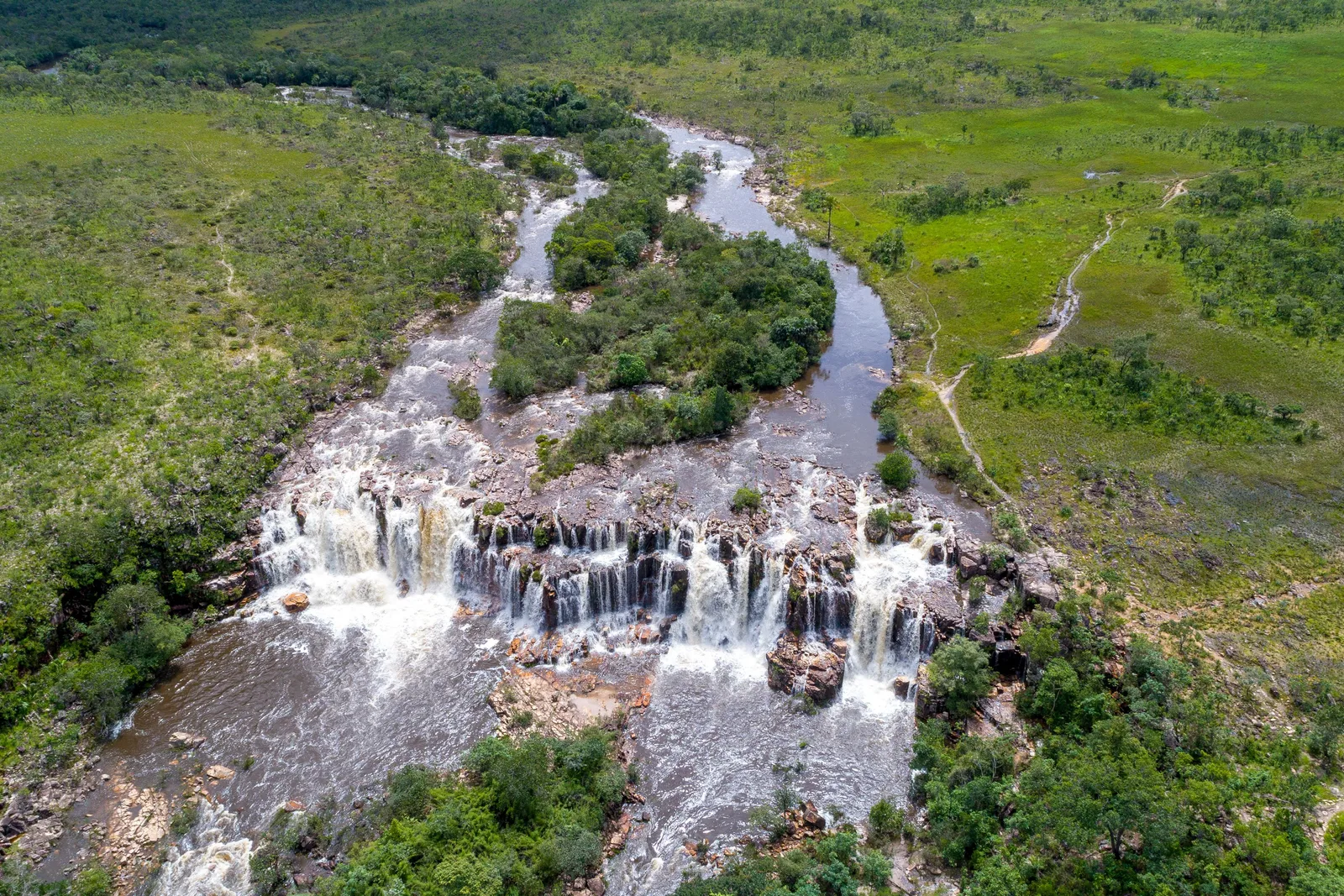 An aerial view of a river splitting around a wooded island and falling over a long ledge of dark rock across green grassland