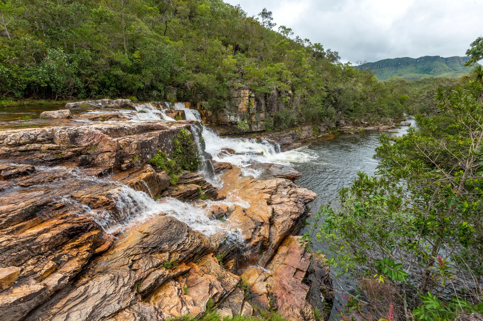 Water running over layered orange rock into a river, with forested slopes and blue hills behind