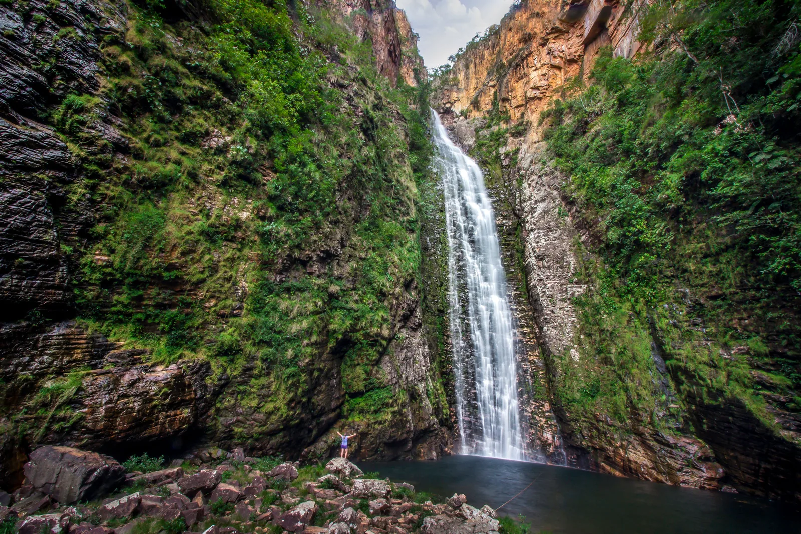 A tall ribbon of water falling down a narrow gorge between mossy dark rock walls into a small pool