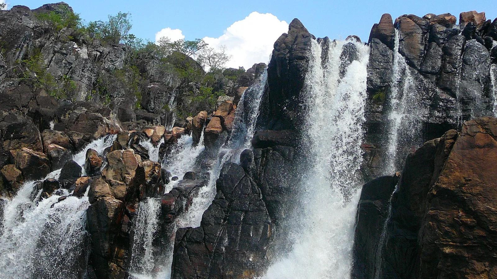 White water pouring in several streams down a wall of dark, jagged rock blocks, with cerrado trees along the top under a blue sky and one white cloud