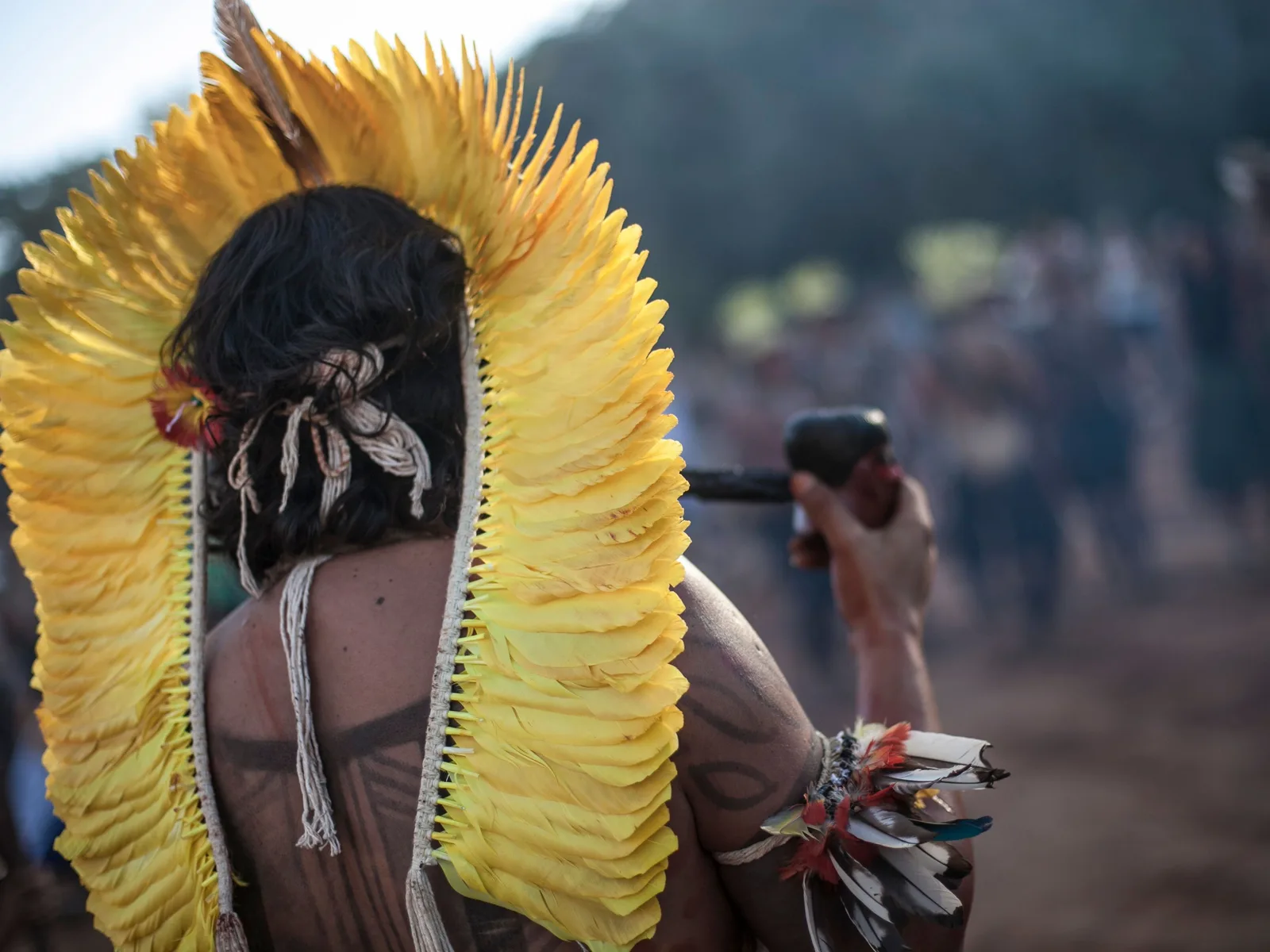 A man seen from behind wearing a tall headdress of yellow feathers, with a blurred crowd on a dirt road ahead of him