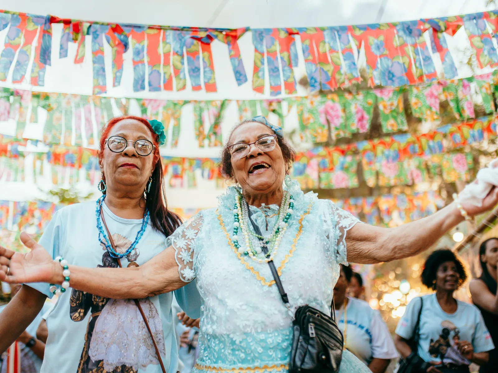 Two older women of a congada group, in light blue, dancing with arms outstretched under strings of colorful paper bunting in a festival procession through São Jorge, a crowd behind them