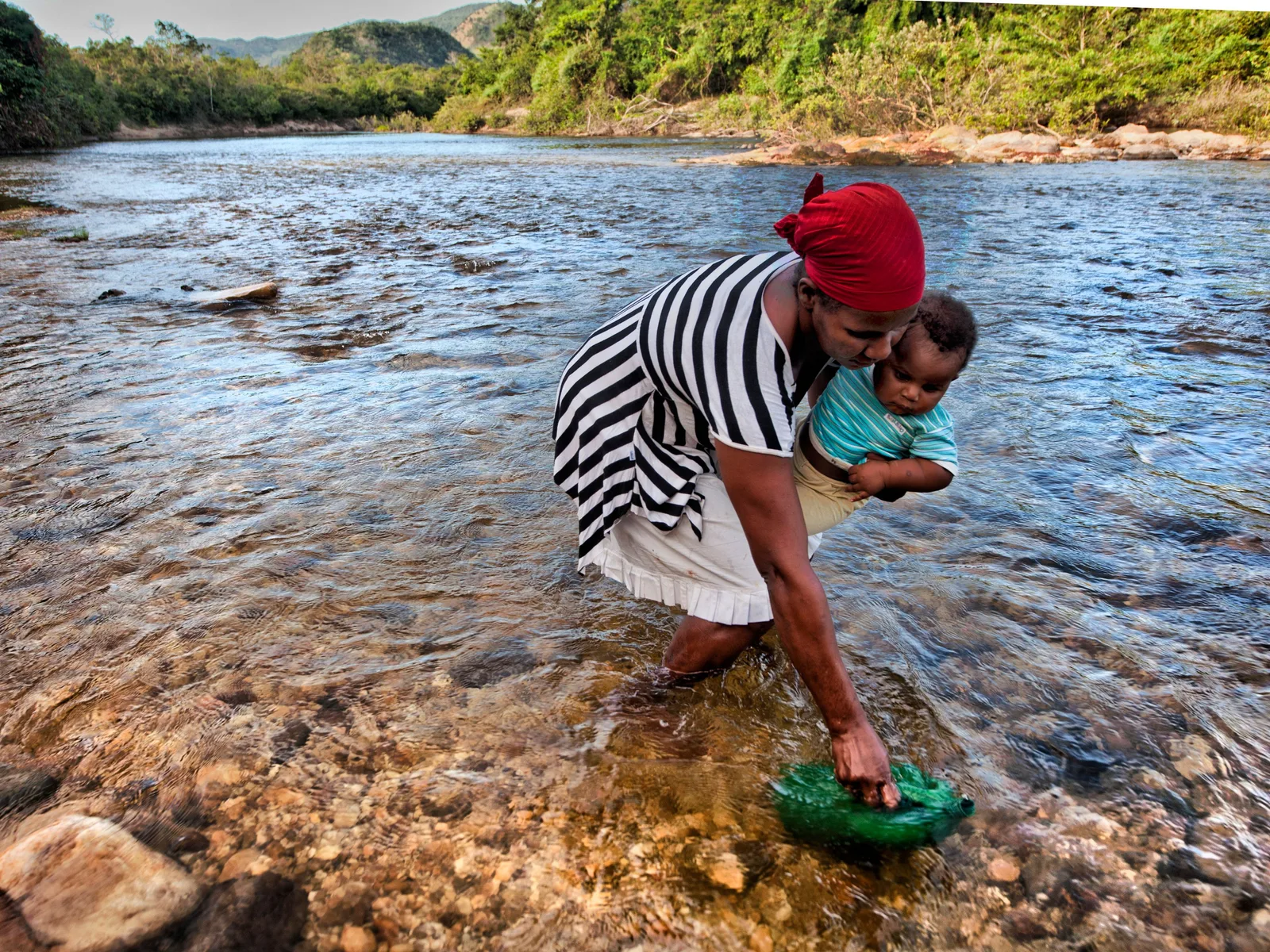 A woman in a red headscarf and striped dress bending to fill a green basin in a clear, shallow river while carrying a baby, wooded hills behind