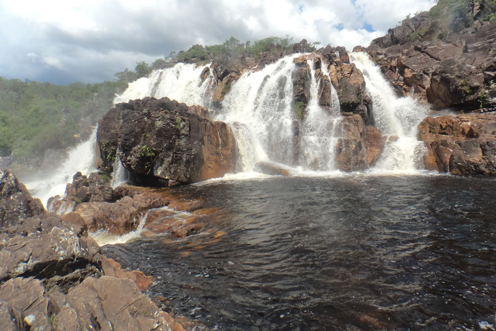 A waterfall spilling over a broad rock ledge into a dark pool, with cerrado on the far bank