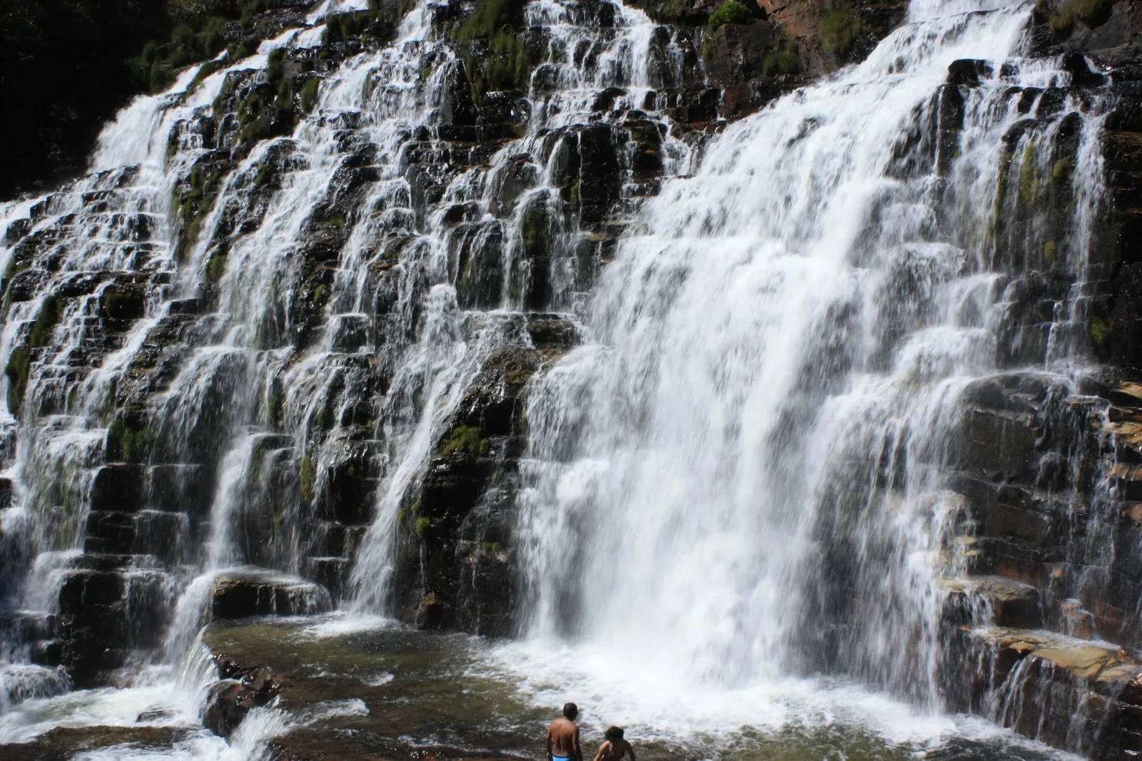 Water pouring over wide steps of dark layered rock, with two people standing in the shallow pool at its foot