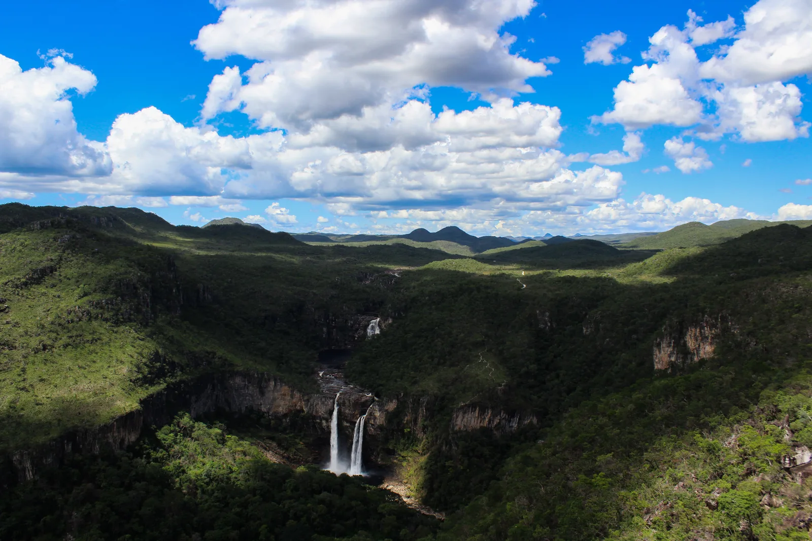A deep green valley under a sky full of white clouds, with a double waterfall far below falling off a pale cliff