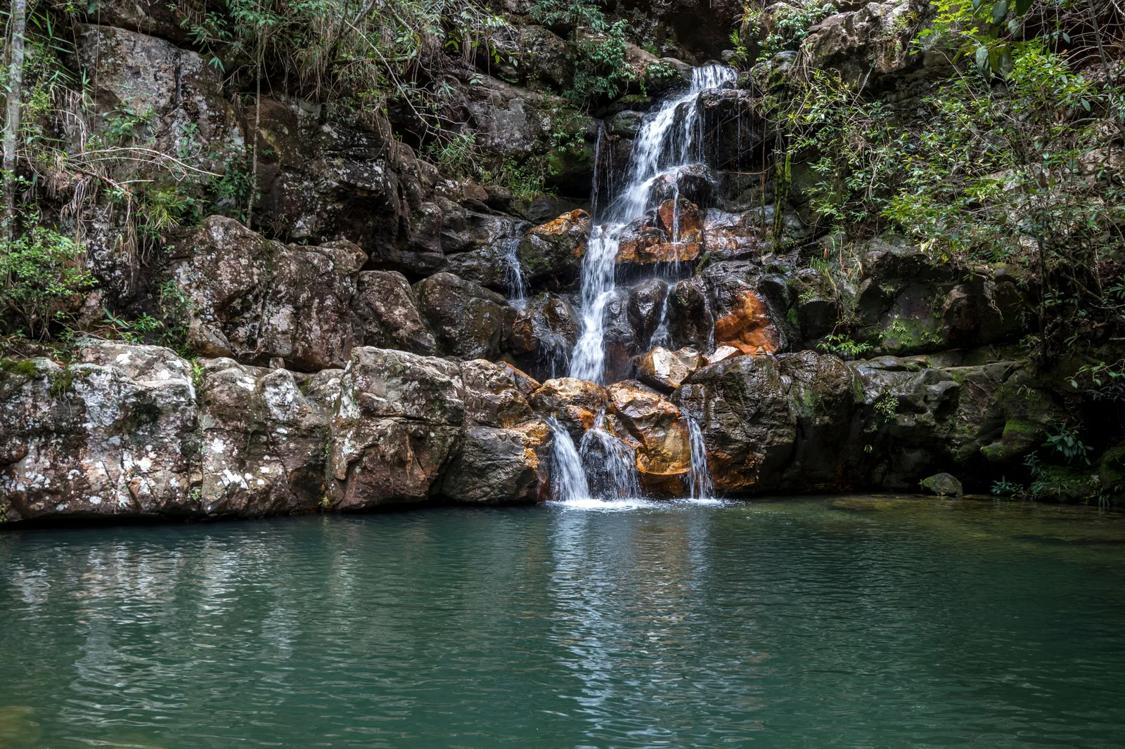 A thin waterfall dropping down a rock face into a wide, clear turquoise pool ringed by pale rock and forest