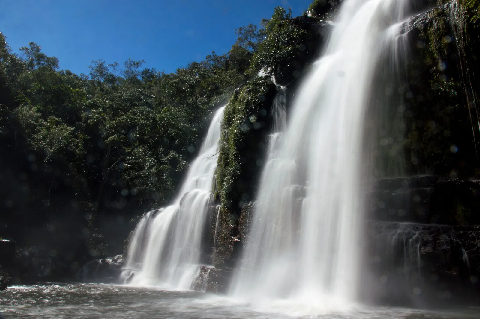 A long-exposure view of a broad waterfall falling in two smooth white curtains into a pool between dark wooded cliffs