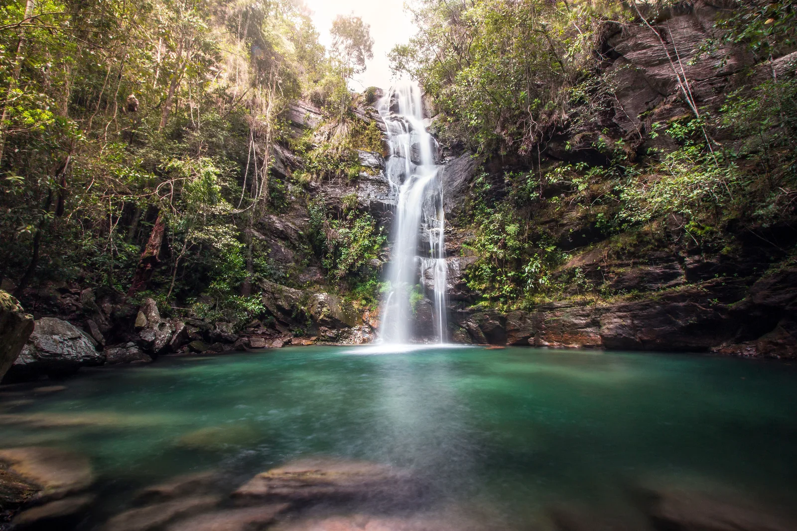 A thin waterfall falling into a turquoise pool surrounded by forest and rock, its surface glassy