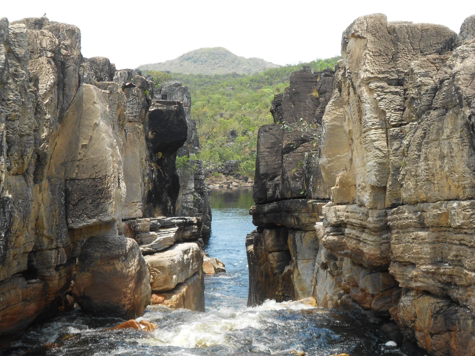 A narrow gap between two tall walls of pale layered rock, with river water rushing through it and a green hill beyond