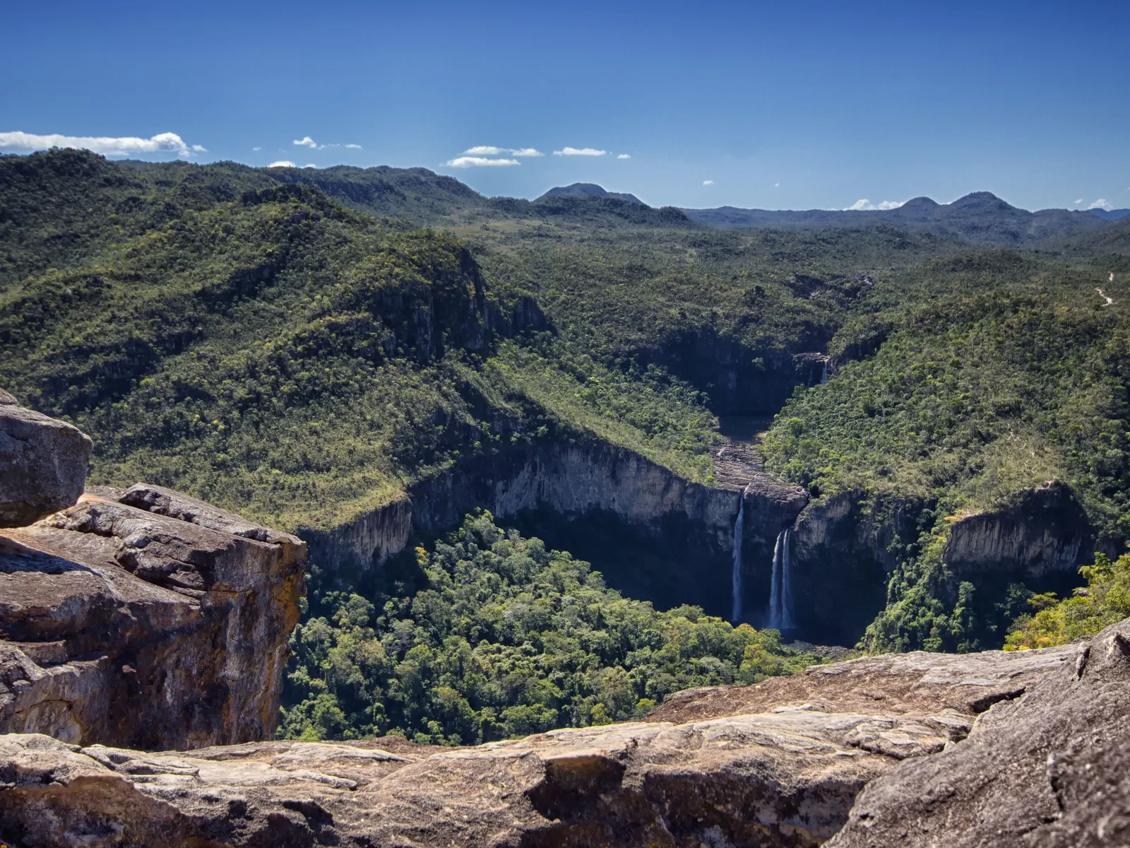 Boulders on a cliff edge in the foreground and, across a deep wooded canyon, two waterfalls dropping off a pale rock wall