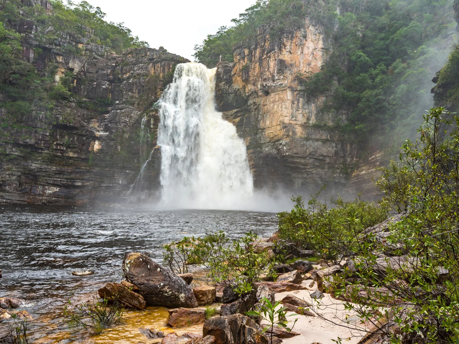 A broad waterfall pouring between tall pale canyon walls into a wide dark pool, with spray drifting over a sandy bank and boulders in front