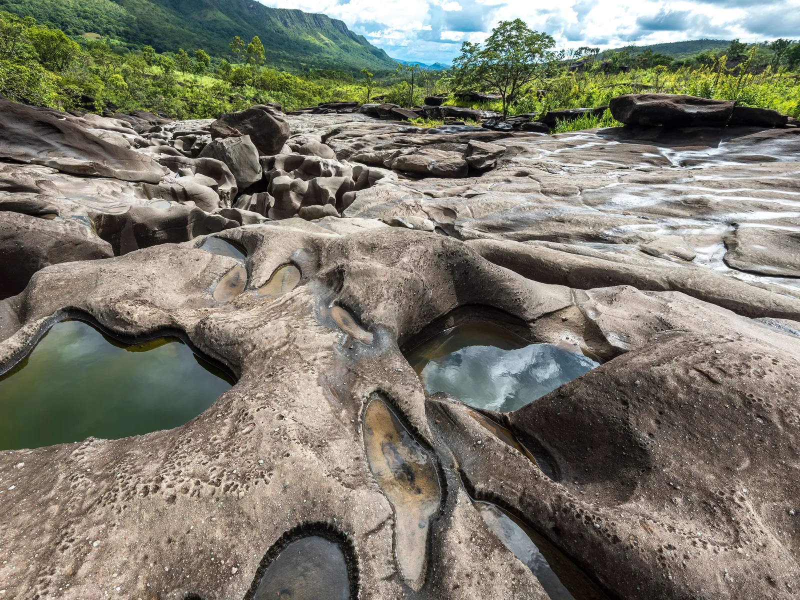 Smooth gray rock riddled with round water-filled basins in the foreground, a river channel behind and wooded hills under a cloudy sky
