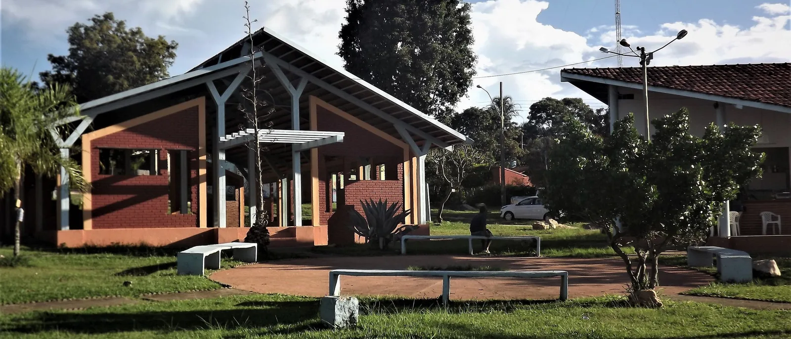 A low pavilion with terracotta walls and a white-framed gabled roof on a grassy community square in Alto Paraíso, with benches in front