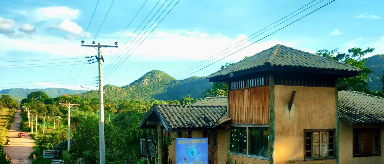 The Casa de Cultura in Cavalcante, a two-story house with earth walls, a wooden upper floor and a tiled roof, beside a dirt road running downhill toward green wooded hills