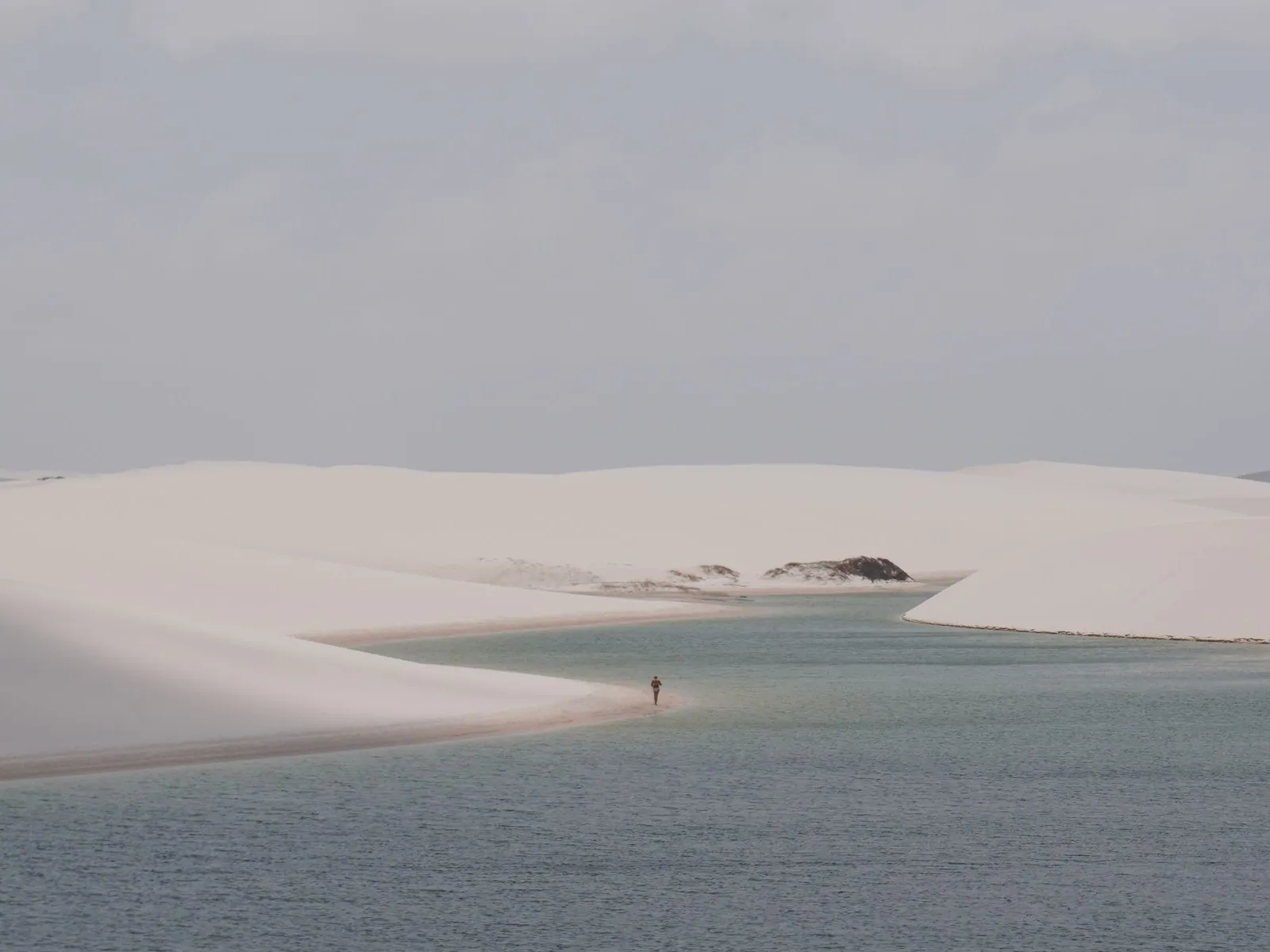 A single tiny figure at the edge of a wide still lagoon ringed by white dunes