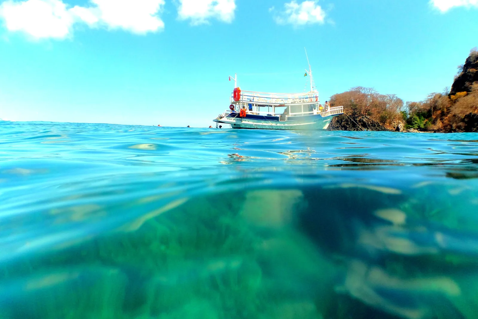 A half-underwater view off Baía do Sancho: clear green water over reef with two snorkelers at the surface, and a white tour boat moored against a rocky shore above them