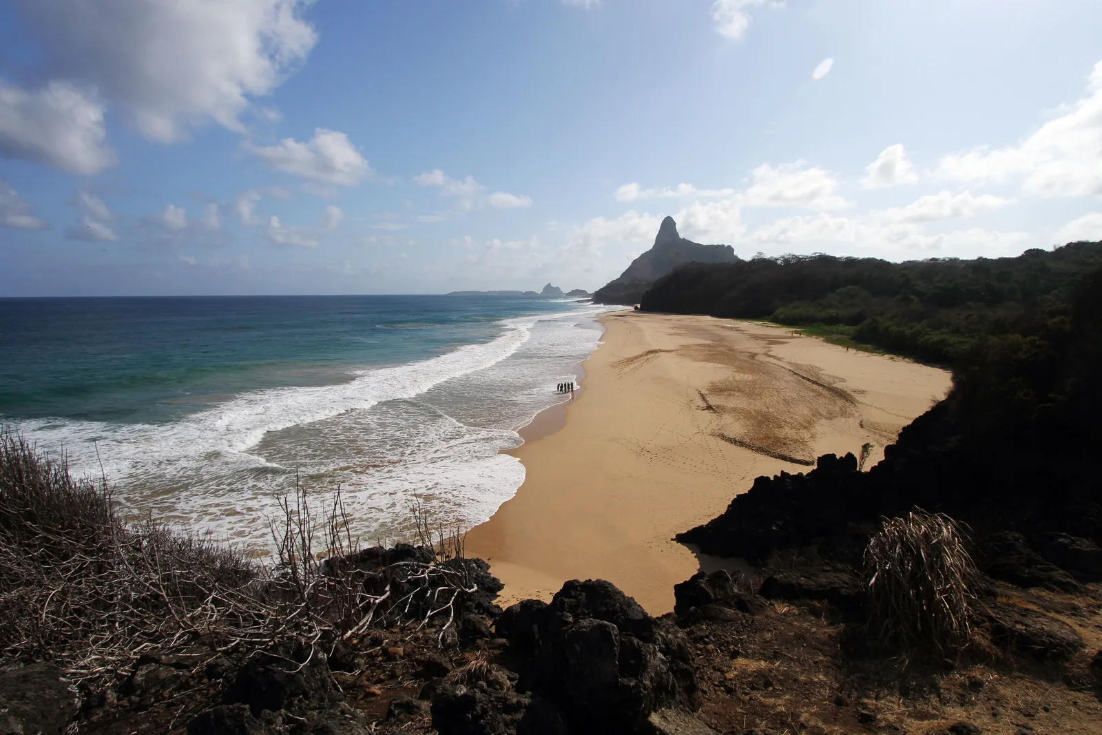The long pale beach at Cacimba do Padre from a rocky rise at one end, surf running up the sand and the pointed silhouette of Morro do Pico in the distance