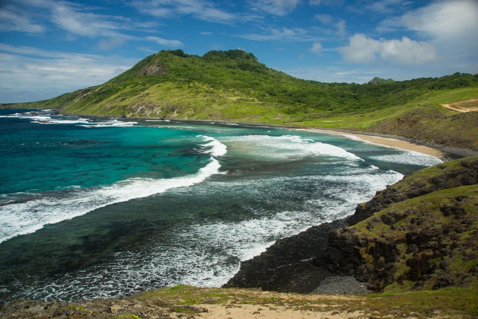 A curved bay backed by steep green hills at Enseada da Caieira, with a pale beach and surf breaking over a wide shallow reef in the foreground