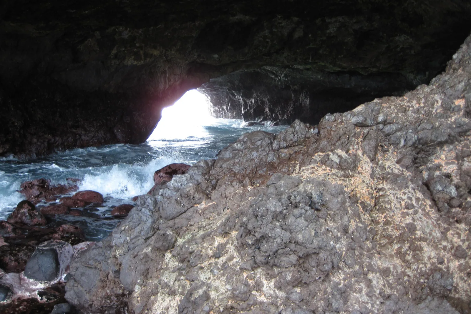 A wide sea cave on the Capim Açu trail, its rock roof arching over a sloping rock shelf with daylight and white surf breaking through the opening beyond