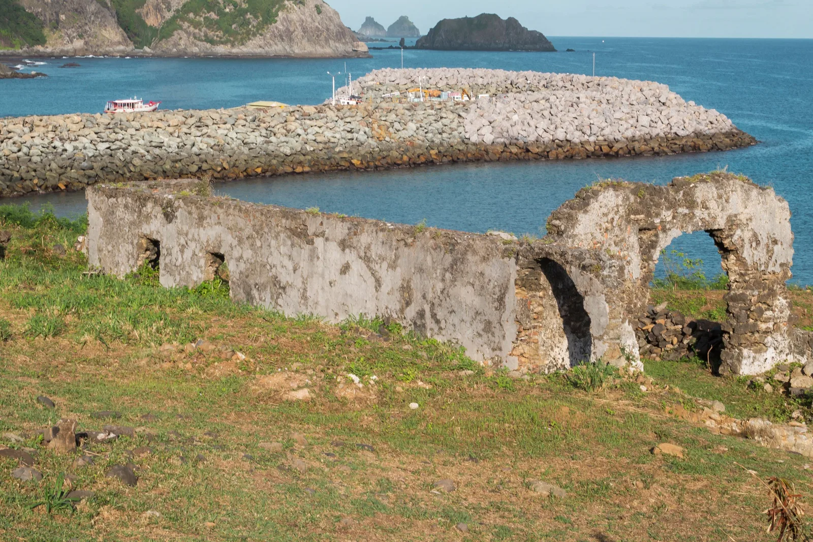The ruined stone arches of the Forte de Santo Antônio standing on rough grass above the harbor, with the long breakwater and calm water behind them