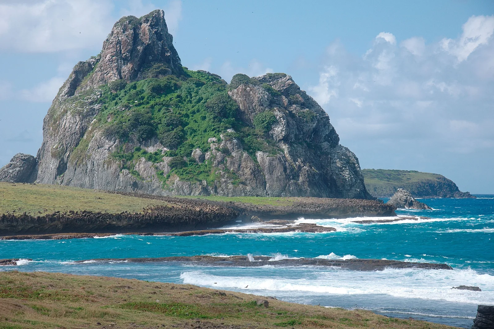 A steep uninhabited rock island of the archipelago rising out of blue water, its top covered in low green vegetation, with a second island behind it