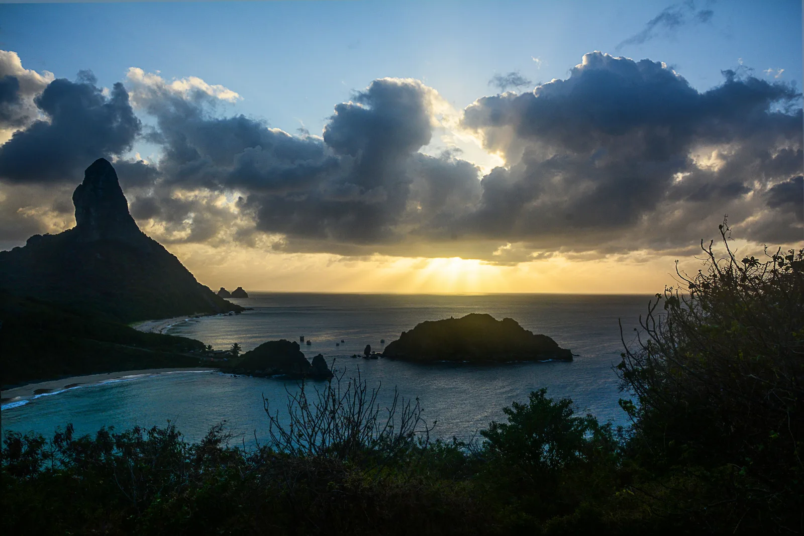 The sun dropping behind heavy cloud over the sea, seen from a clifftop viewpoint with the pointed silhouette of Morro do Pico on the left and a rock island below