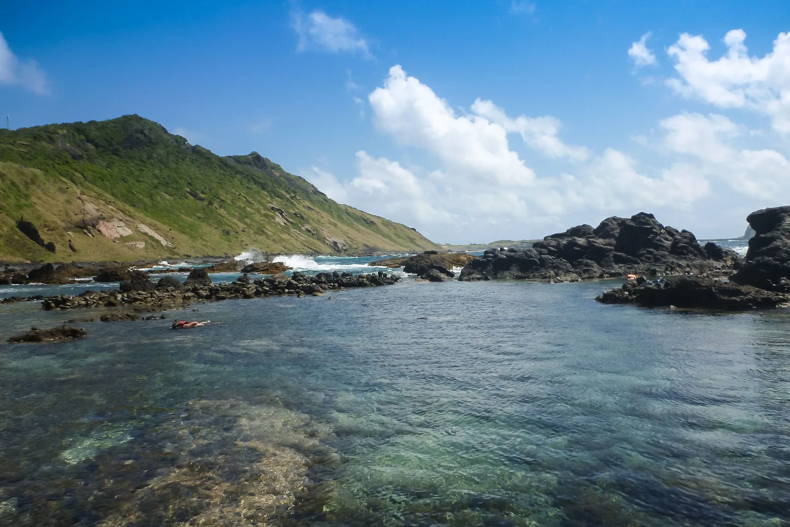 A shallow, glass-clear pool inside a low rocky shore with one snorkeler floating face down in it, a green and tan hillside running down on the left and surf breaking on the reef beyond