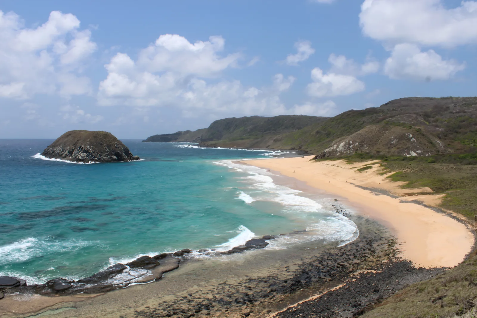 A wide empty beach at Praia do Leão curving between green headlands, with a dark rock island standing offshore and turquoise water breaking over a shallow reef