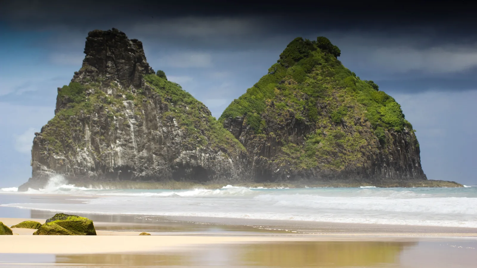 A long exposure of Morro Dois Irmãos from the beach under a dark storm sky, the twin peaks lit and green against it, and the water smoothed to a pale sheet across the sand